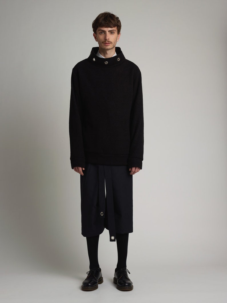 May include: A black mock neck sweater with a high collar and silver button accents. The sweater is paired with black pants and black shoes.