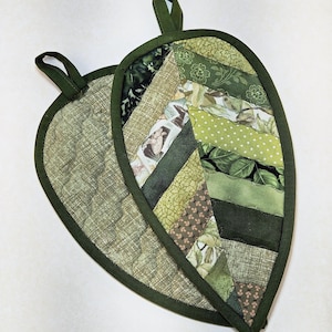 May include: Two leaf-shaped quilted pot holders. The pot holders are primarily green, with a variety of green fabric patterns, including floral, polka dot, and solid shades. Each has a dark green binding and a loop for hanging.