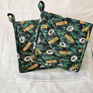 May include: Two green and gold quilted pot holders with the word "Packers" repeated on them. The pot holders are sitting on a clear glass baking dish.