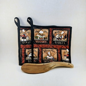 May include: A quilted pot holder with black trim and loops, featuring six square panels with chef illustrations and text. A wooden spoon rests in front of the pot holder. The panels depict various food categories like seafood, veggies, and barista.