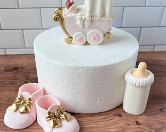 May include: A baby shower cake topper set featuring a pink baby carriage with a baby, pink baby shoes with gold bows, and a white baby bottle with a yellow teat. The carriage has gold accents and flower details.