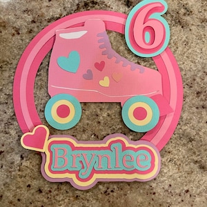 May include: A pink and blue paper craft with a roller skate, a heart, and the name "Brynnlee" with the number "6" in the background.