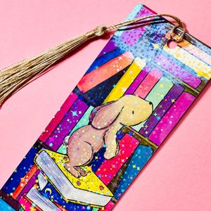 May include: A colourful bookmark with a glittery finish features a cartoon bunny rabbit standing on a stack of books. The bookmark has a gold tassel.