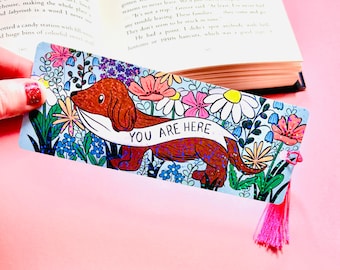 Puede incluir: Un marcapáginas colorido con una ilustración de dachshund y flores. El marcapáginas tiene un borla rosa y el texto "YOU ARE HERE" en una pancarta blanca.