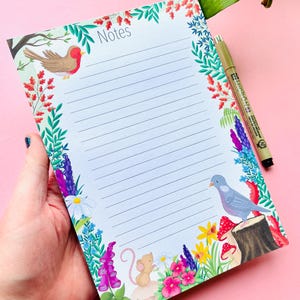 May include: A notepad with the word "Notes" at the top, surrounded by a floral and fauna border. The border features illustrations of birds, flowers, mushrooms, and a mouse. A black pen rests on the notepad. The notepad is held by a hand.