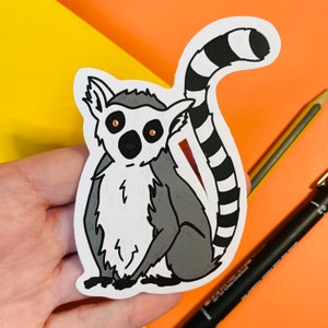 May include: A black and white sticker of a lemur with a ringed tail. The lemur is sitting on its haunches with its arms crossed. The lemur has large, round eyes and a small, pointed nose.
