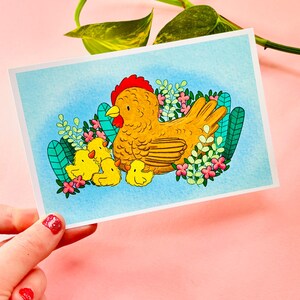 May include: A watercolour illustration of a mother hen with three chicks. The hen is brown with a red comb and the chicks are yellow. The illustration is set against a blue background with green leaves and pink flowers.