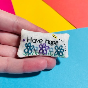 May include: White fabric cushion with embroidered flowers and the text "Have hope".