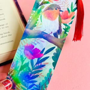 Puede incluir: Marcapáginas decorativo con una ilustración en acuarela de un petirrojo posado en una rama, rodeado de flores y follaje. El marcapáginas tiene una borla roja. La paleta de colores incluye verdes, rosas y azules.