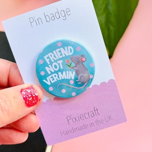 Könnte beinhalten: Ein runder Ansteck-Pin mit dem Text "FRIEND NOT VERMIN" auf hellblauem Hintergrund mit rosa Tupfen. Der Pin zeigt eine graue Maus, die eine Blume hält. Der Pin befindet sich auf einer weißen Karte mit der Aufschrift "Pin badge".