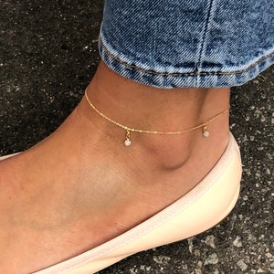 Könnte beinhalten: Ein zartes goldenes Fußkettchen mit kleinen, baumelnden Anhängern. Das Fußkettchen wird an einem Knöchel mit einem hellrosa Ballettschuh und einer blauen Jeans im Hintergrund getragen. Es ist ein schlichter, eleganter Schmuck.
