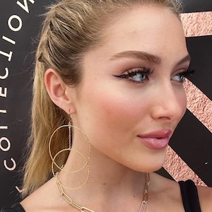 Puede incluir: Una mujer con un top negro y un collar de cadena dorada con un colgante negro y plateado. Lleva pendientes de aro dorados con un acabado texturizado.