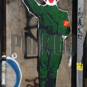 Puede incluir: Una plantilla verde y negra de un payaso con pelo rojo y una 'M' amarilla en su uniforme, haciendo un saludo. El fondo es una pared de hormigón gris con grafitis.