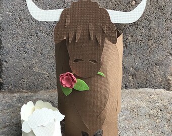 May include: A brown papercraft Highland cow with a white horn and a pink flower, standing next to a white papercraft sheep.