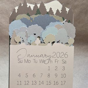Pode incluir: Um calendário de papel para janeiro de 2026, com uma cena de floresta em camadas em tons de verde, cinza e bege. O calendário é suportado por um suporte branco. O mês e o ano estão escritos em itálico.