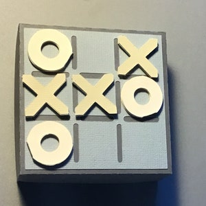 May include: A handmade tic-tac-toe game. The game board is light blue with a gray border. The X's and O's are cream-colored and placed on the board. The game is set on a gray surface.