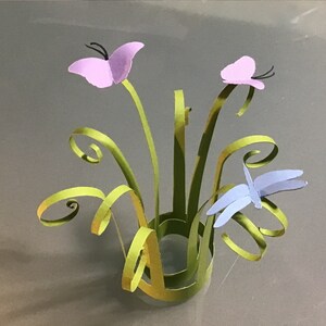Puede incluir: Un arreglo floral de papel con dos mariposas lavanda y una libélula azul claro. El arreglo incluye tallos verdes y hojas rizadas, todo sobre un fondo gris. Las mariposas tienen antenas negras.