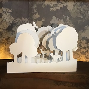Op de afbeelding: Witte papieren uitsnijding van een bos scène met bomen en herten. De scène is gelaagd om een 3D-effect te creëren.