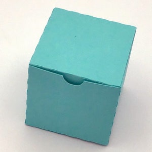 Könnte beinhalten: Eine kleine, quadratische, türkisfarbene Geschenkbox mit einem gebogenen Ausschnitt auf der vorderen Klappe. Die Box besteht aus dickem Papier oder Karton und hat sichtbare Nähte an den Kanten. Die Box ist leicht angewinkelt.