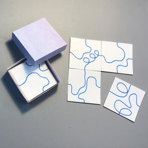 May include: A set of white square coasters with blue abstract line art, displayed with a matching lavender box. The coasters are arranged on a gray surface, with some inside and outside the box.
