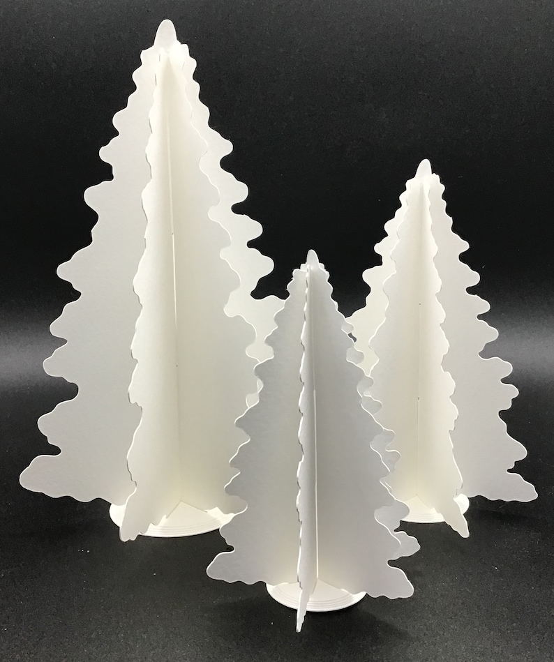 May include: Three white paper Christmas trees of varying sizes. The trees are made of folded paper and have a scalloped edge. The trees are standing on a black surface.