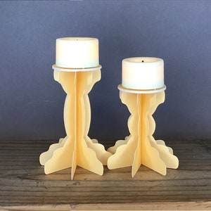 Könnte beinhalten: Zwei gelbe Kerzenhalter mit weißen Kerzen. Die Kerzenhalter sind aus Pappe und haben ein gewelltes Design.