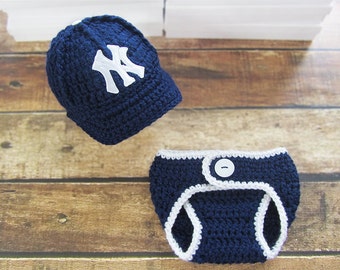 Puede incluir: Conjunto de bebé de ganchillo azul marino, que incluye una gorra de béisbol con el logotipo "NY" blanco y una braguita con ribete blanco. El conjunto está hecho a mano y tiene un cierre de botón. La palabra "Navy" está impresa en la parte inferior.