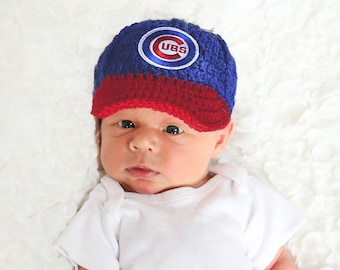 Puede incluir: Gorra de béisbol azul y roja de ganchillo con el logotipo de los Chicago Cubs. La gorra la lleva un bebé con un body blanco.