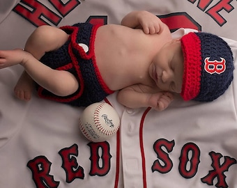 Puede incluir: Un bebé recién nacido con gorra y cubrepañales de béisbol azul marino y rojo, acostado sobre una camiseta de béisbol blanca con "RED SOX" en letras rojas. Una pelota de béisbol descansa cerca del bebé.