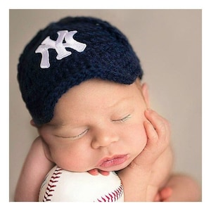 Puede incluir: Un beb&eacute; reci&eacute;n nacido durmiendo, con un gorro de punto azul marino con el logo blanco "NY", apoyado en una pelota de b&eacute;isbol. El beb&eacute; est&aacute; dormido, con la mejilla apoyada en la mano, y la pelota de b&eacute;isbol en primer plano.