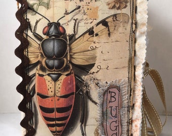 Könnte beinhalten: Ein handgemachtes Journal mit einem braunen Stoffbezug, einer Spitzenborte und einer Vintage-Insektenillustration. Der Umschlag zeigt ein braunes und rotes Insekt mit schwarzem Kopf und Flügeln. Das Journal hat einen braunen Bandriemen und das Wort "BUGS" ist auf einem braunen Etikett geschrieben.
