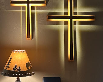 May include: Two illuminated wooden crosses with a dark border are mounted on a wall. A lamp with a pinecone base and a shade featuring a forest scene sits below. A blue book with the words "Holy Bible" is also present.