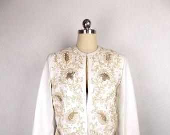 May include: A white jacket with a gold and brown floral embroidery design. The jacket has a mandarin collar and long sleeves.