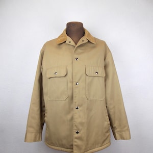 May include: Tan work jacket with a corduroy collar, two chest pockets, and silver snap closures. The jacket is displayed on a mannequin. A classic, durable outerwear piece.