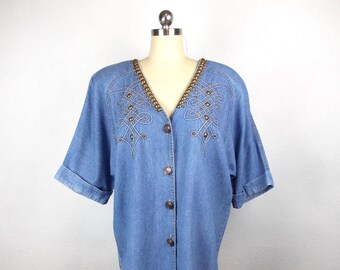 May include: A light blue denim button-down shirt with short sleeves. The shirt features decorative gold-colored beading along the V-neckline and embroidered patterns on the front. The shirt is displayed on a mannequin.