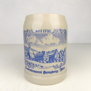 May include: A white ceramic beer stein with blue and white illustrations. The stein features a depiction of a building with the text "Klosterbrauerei Kreuzberg/Rhön" and "seit 1731".