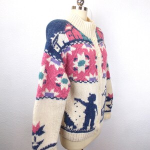 May include: Cream-colored, button-up cardigan sweater with a high collar. The sweater features a colorful, patterned design with pink, blue, purple, and teal accents. A silhouette of a person and dog is on the lower portion of the sweater.