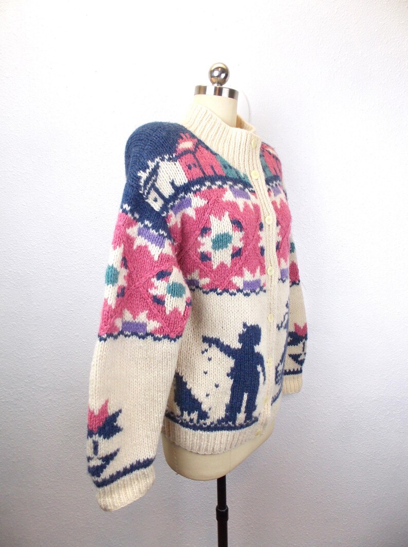 May include: Cream-colored, button-up cardigan sweater with a high collar. The sweater features a colorful, patterned design with pink, blue, purple, and teal accents. A silhouette of a person and dog is on the lower portion of the sweater.