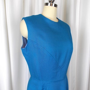 May include: A sleeveless, teal blue dress displayed on a mannequin. The dress features a round neckline and a fitted bodice with a subtle V-shaped seam detail. The skirt flares out from the waist, creating a classic silhouette.