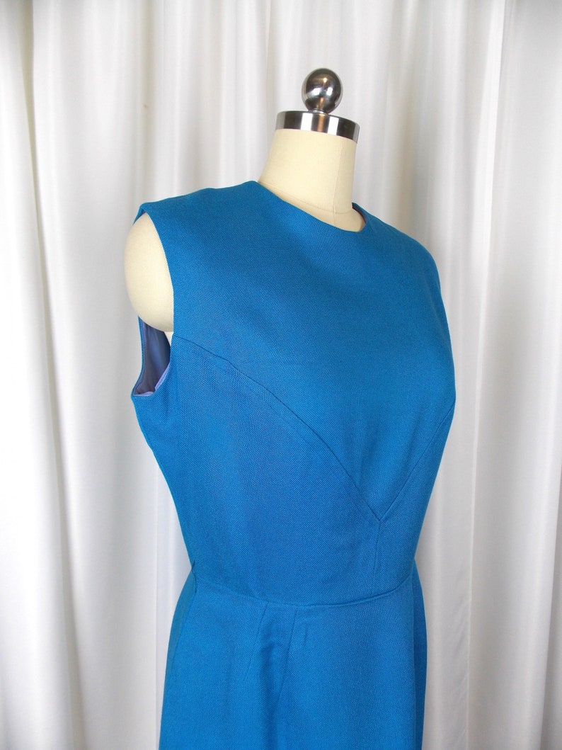 May include: A sleeveless, teal blue dress displayed on a mannequin. The dress features a round neckline and a fitted bodice with a subtle V-shaped seam detail. The skirt flares out from the waist, creating a classic silhouette.