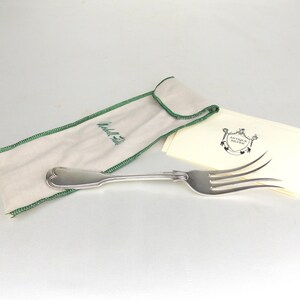 May include: A silver serving fork with a decorative handle, alongside a cream-coloured pouch with green stitching and text. A stack of cream-coloured cards with a crest logo is also present. The items are on a white surface.