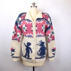 May include: Cream-colored, button-down cardigan sweater with a collared neckline. The sweater features a geometric pattern in pink, blue, and purple. The lower portion of the sweater has silhouettes of a boy and girl. The sleeves have a star design.