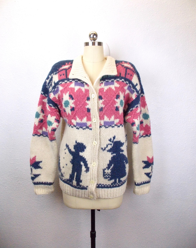 May include: Cream-colored, button-down cardigan sweater with a collared neckline. The sweater features a geometric pattern in pink, blue, and purple. The lower portion of the sweater has silhouettes of a boy and girl. The sleeves have a star design.