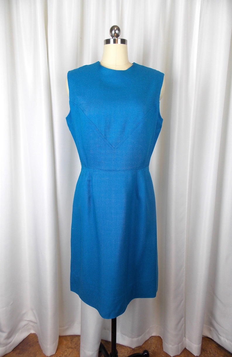 May include: A sleeveless, knee-length teal dress displayed on a mannequin. The dress features a V-neckline and a fitted waist. The fabric has a subtle texture, and the dress is a solid, vibrant color.