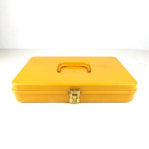 May include: A bright yellow rectangular toolbox with a handle on top and a gold-colored clasp. The toolbox is closed and sits on a white surface. The handle is a curved, recessed design.