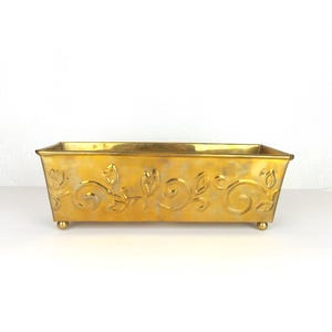 May include: A rectangular, gold-colored metal planter with floral and swirl designs. The planter sits on four small, rounded feet. The surface has a slightly aged appearance, adding to its vintage charm.
