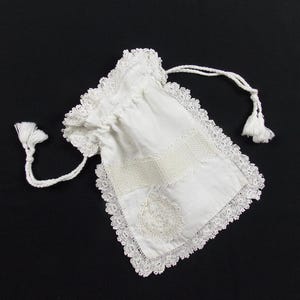 May include: A white fabric drawstring pouch featuring intricate lace trim along the edges, a horizontal band of decorative openwork stitching across the center, and a circular lace appliqué detail on the lower portion, finished with two white tassels.