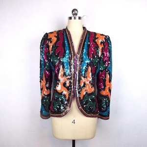 May include: A colorful sequined jacket with a V-neck design. The jacket features a vibrant pattern of orange, teal, green, and magenta sequins. The jacket is displayed on a mannequin.