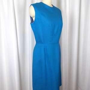 May include: A sleeveless, knee-length teal dress displayed on a mannequin. The dress features a fitted bodice with a subtle V-neckline and a defined waist. The fabric appears to be a textured weave. The dress is a solid color and has a classic, vintage style.