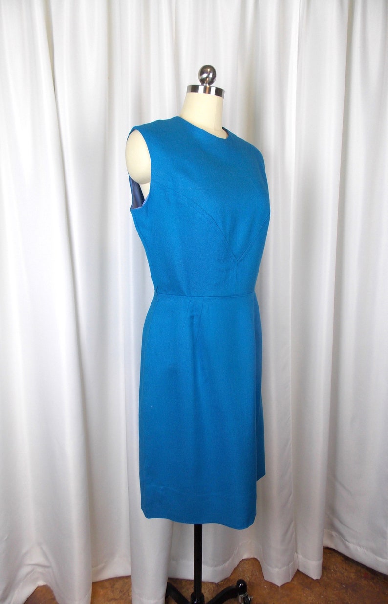 May include: A sleeveless, knee-length teal dress displayed on a mannequin. The dress features a fitted bodice with a subtle V-neckline and a defined waist. The fabric appears to be a textured weave. The dress is a solid color and has a classic, vintage style.